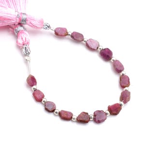 Può includere: Una collana di pietre preziose grezze, rosa-rossastre, con perline argentate. Le pietre preziose sono infilate insieme e la collana è attaccata a un nappa rosa con accenti argentati. Le pietre preziose sono di dimensioni e forme variabili.