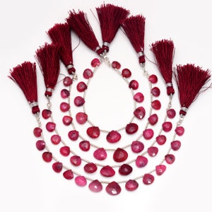 May include: Several strands of heart-shaped, deep red gemstones are strung on silver wire. Each strand is finished with a burgundy tassel. The gemstones vary in shade from light to dark red. The arrangement is on a white background.