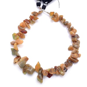 Könnte beinhalten: Eine Halskette aus unregelmäßig geformten, polierten Edelsteinen in Orange-, Braun- und Grüntönen. Die Steine sind mit einem silberfarbenen Verschluss und einer schwarzen Quaste aufgereiht. Die Halskette wird vor weißem Hintergrund präsentiert.