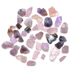 May include: A selection of raw gemstones in shades of purple, pink, and grey. The stones are scattered on a white surface, displaying their natural textures and shapes. Some stones are translucent, while others are opaque, offering a variety of colours.