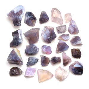 May include: A collection of raw, uncut gemstones in various shapes and sizes. The stones display a range of colors, including shades of purple, lavender, gray, and hints of pink and brown. The gemstones are arranged on a white surface.