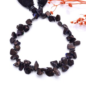 Könnte beinhalten: Eine Halskette aus dunklen, unregelmäßig geformten Perlen mit schillernden Farbtönen. Die Perlen sind zu einer kreisförmigen Form aufgefädelt. Schwarze Quasten sind oben sichtbar. Bei den Perlen handelt es sich wahrscheinlich um Edelsteine oder polierte Steine.