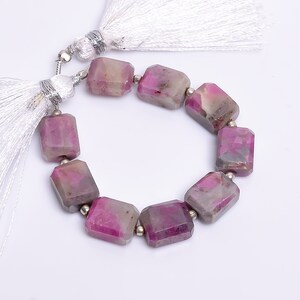 Puede incluir: Un collar de piedras preciosas rectangulares rosas y grises, cada una facetada. Pequeñas cuentas plateadas separan las piedras. Borlas blancas están unidas a cada extremo. Las piedras están dispuestas en un patrón circular.