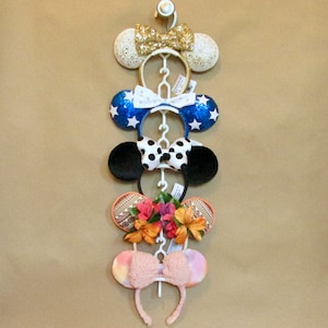 May include: Five decorative headbands with mouse ears and bows, displayed on a white rack. The headbands have different designs: gold glitter, blue with white stars, black with white polka dots, floral, and pink. The rack is against a neutral background.