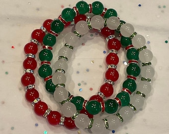 Puede incluir: Tres pulseras de cuentas rojas, verdes y blancas con detalles plateados. Las pulseras están hechas de cuentas redondas y están apiladas una encima de la otra.