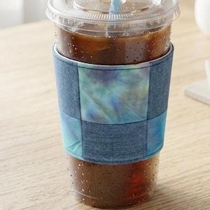 May include: A clear plastic iced coffee cup with a light blue straw and a fabric sleeve featuring a patchwork design of blue denim squares and tie-dye squares in shades of teal, purple, and blue, wrapped around the center of the cup.