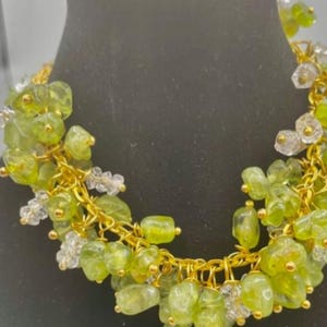 May include: A gold chain necklace featuring clusters of light green and clear gemstones. The gemstones are irregularly shaped and clustered, creating a unique design. The necklace is displayed on a black surface, showcasing the jewellery's details.
