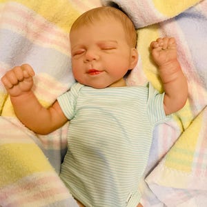Puede incluir: Una mu&ntilde;eca beb&eacute; realista con un body a rayas azul claro y blanco, acostada sobre una manta colorida. La mu&ntilde;eca tiene los ojos cerrados, la boca ligeramente abierta y los pu&ntilde;os cerrados. La manta tiene rayas pastel amarillas, rosas y azules.
