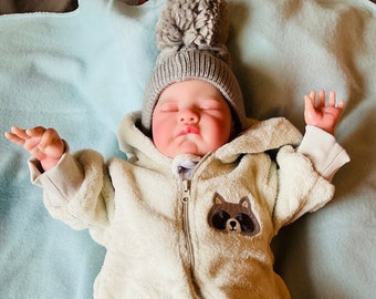 Puede incluir: Un bebé con un gorro gris con pompón y un mono crema con cremallera y un parche de mapache. El bebé también lleva calcetines a rayas azules y blancas con un diseño de perro. El bebé está acostado sobre una manta azul claro.