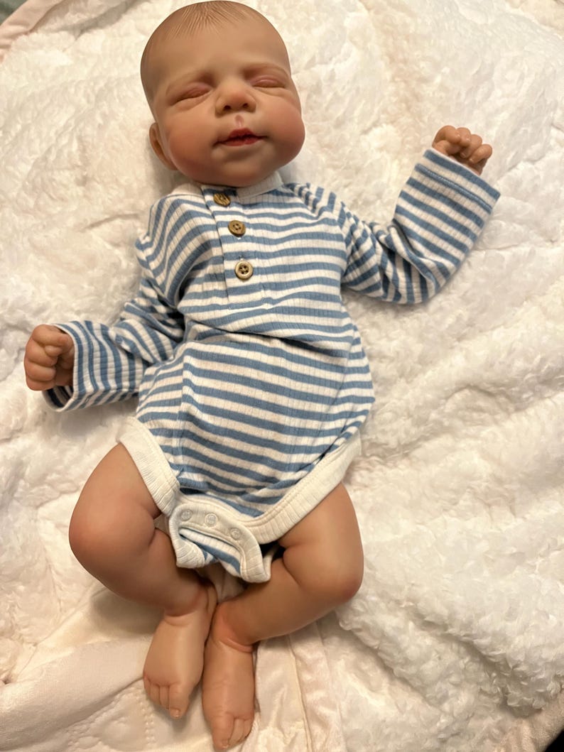Puede incluir: Una mu&ntilde;eca beb&eacute; realista con los ojos cerrados, acostada sobre una manta blanca y esponjosa. La mu&ntilde;eca lleva un body a rayas azules y blancas con botones de madera. La piel de la mu&ntilde;eca tiene un tono realista.
