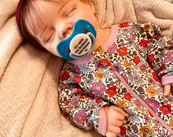 May include: A sleeping baby doll wearing a dark burgundy flower headband and a floral print onesie with a zipper. The doll has a blue pacifier in its mouth with the text 50% Wild, 50% Brilliant, 100% Yours printed on the center of the pacifier.