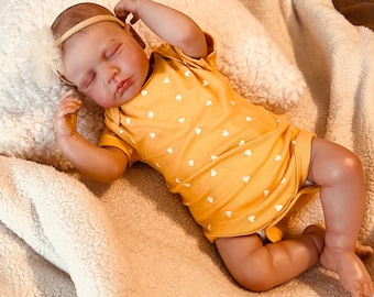 Puede incluir: Un muñeco de bebé durmiendo que lleva un mono amarillo con corazones blancos impresos. El muñeco está tumbado sobre una manta blanca y esponjosa.