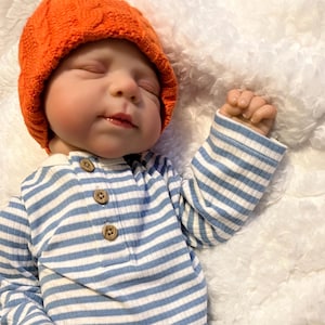 Puede incluir: Una mu&ntilde;eca beb&eacute; con un gorro naranja tejido y una camisa de manga larga a rayas azules y blancas. La mu&ntilde;eca tiene los ojos cerrados y una expresi&oacute;n tranquila. La camisa tiene tres botones de madera.