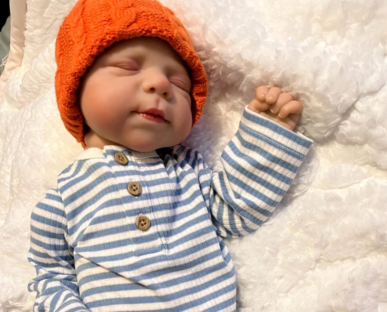 Puede incluir: Una mu&ntilde;eca beb&eacute; con un gorro naranja tejido y una camisa de manga larga a rayas azules y blancas. La mu&ntilde;eca tiene los ojos cerrados y una expresi&oacute;n tranquila. La camisa tiene tres botones de madera.