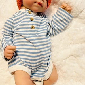 Puede incluir: Un beb&eacute; con un gorro de calabaza de punto naranja y un body a rayas azules y blancas con botones de madera. El beb&eacute; est&aacute; acostado sobre una manta blanca y esponjosa.
