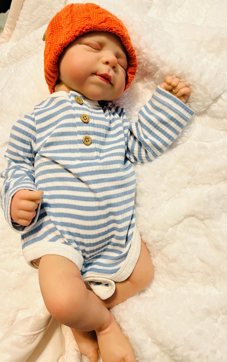 Puede incluir: Un beb&eacute; con un gorro de calabaza de punto naranja y un body a rayas azules y blancas con botones de madera. El beb&eacute; est&aacute; acostado sobre una manta blanca y esponjosa.