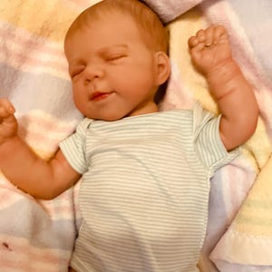 Puede incluir: Una mu&ntilde;eca beb&eacute; realista con los ojos cerrados y una expresi&oacute;n serena. La mu&ntilde;eca lleva un body a rayas verdes y blancas. Los brazos de la mu&ntilde;eca est&aacute;n levantados, descansando sobre una manta suave estampada.