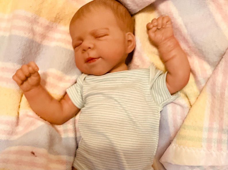 Puede incluir: Una mu&ntilde;eca beb&eacute; realista con los ojos cerrados y una expresi&oacute;n serena. La mu&ntilde;eca lleva un body a rayas verdes y blancas. Los brazos de la mu&ntilde;eca est&aacute;n levantados, descansando sobre una manta suave estampada.