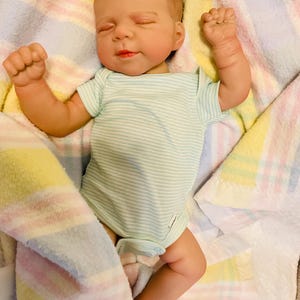 Puede incluir: Una mu&ntilde;eca beb&eacute; realista con un body a rayas azul claro y blanco, acostada sobre una manta colorida. La mu&ntilde;eca tiene los ojos cerrados, la boca ligeramente abierta y los pu&ntilde;os cerrados. La manta tiene rayas pastel amarillas, rosas y azules.