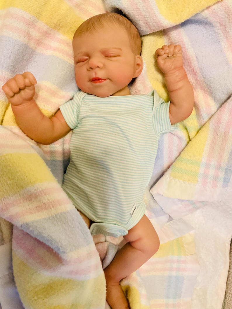 Puede incluir: Una mu&ntilde;eca beb&eacute; realista con un body a rayas azul claro y blanco, acostada sobre una manta colorida. La mu&ntilde;eca tiene los ojos cerrados, la boca ligeramente abierta y los pu&ntilde;os cerrados. La manta tiene rayas pastel amarillas, rosas y azules.