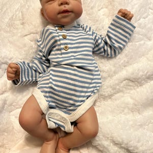 Puede incluir: Una mu&ntilde;eca beb&eacute; realista con los ojos cerrados, vestida con un body a rayas azules y blancas con botones de madera. La mu&ntilde;eca est&aacute; acostada sobre una manta suave y blanca con textura. La piel del beb&eacute; tiene una coloraci&oacute;n realista.
