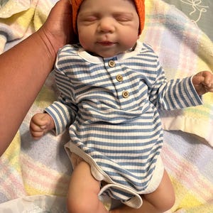 Puede incluir: Una mu&ntilde;eca beb&eacute; realista con un body a rayas azules y blancas con botones de madera y un gorro naranja. La mu&ntilde;eca est&aacute; acostada sobre una manta estampada, con una mano que la sostiene suavemente.