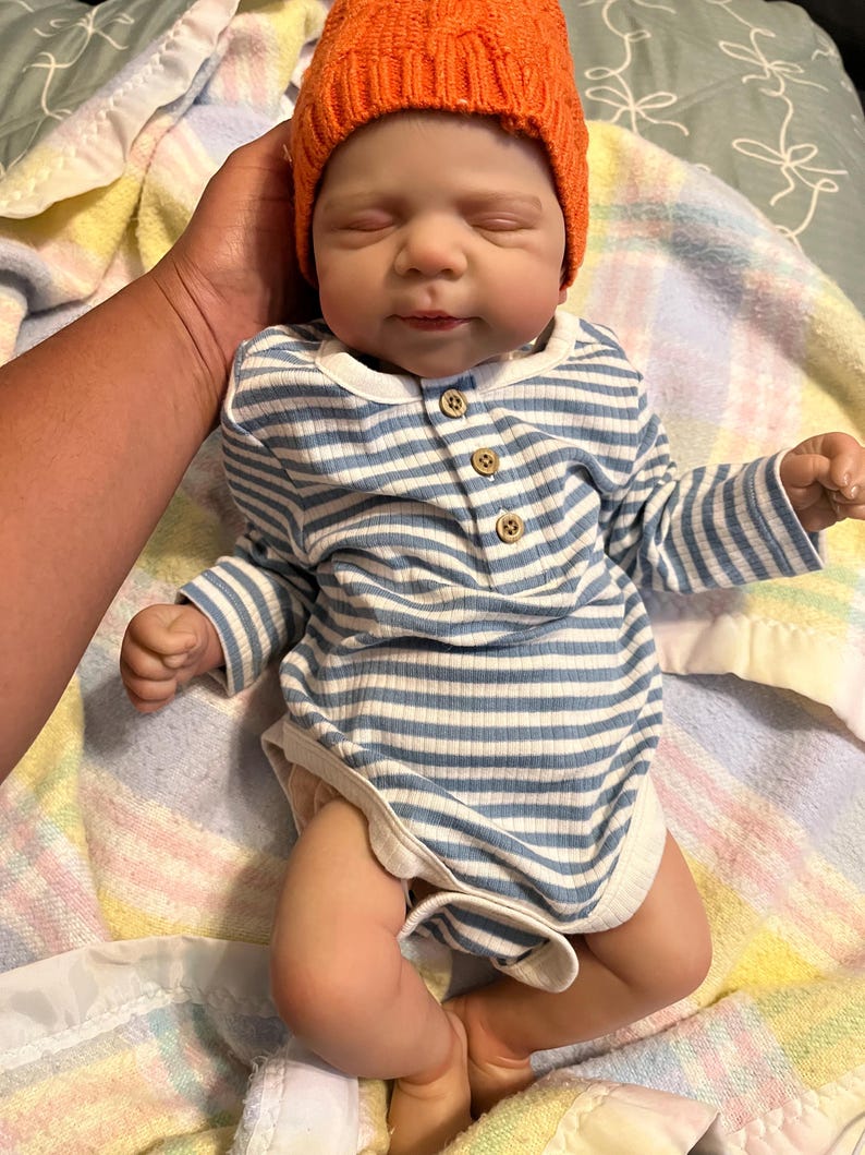 Puede incluir: Una mu&ntilde;eca beb&eacute; realista con un body a rayas azules y blancas con botones de madera y un gorro naranja. La mu&ntilde;eca est&aacute; acostada sobre una manta estampada, con una mano que la sostiene suavemente.
