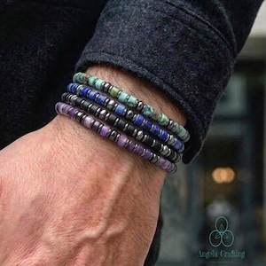 May include: A set of four stacked bracelets featuring small, cylindrical beads in shades of green, blue, black, and purple. Each bracelet is constructed with alternating colored beads and small, metallic silver-toned spacer beads throughout.