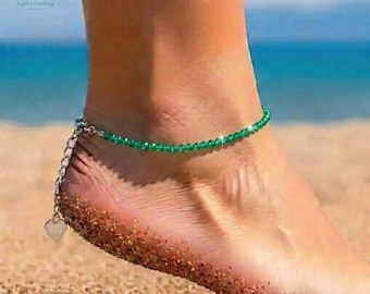 May include: A green beaded anklet with a silver chain and heart charm. The anklet is worn on an ankle on a sandy beach with the ocean in the background. The anklet is made of small, round green beads.