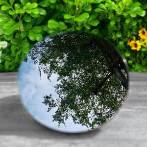 May include: A circular, polished glass sphere or paperweight featuring a reflective surface that displays a clear, inverted image of a tree with green leaves against a light blue sky with soft white clouds, creating a detailed optical effect.
