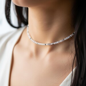 May include: Delicate choker necklace featuring small, round, white beads interspersed with tiny black beads. The necklace is worn around the neck, creating a subtle and elegant accessory. The beads appear to be evenly spaced, creating a refined look.
