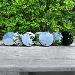 May include: A row of seven circular, reflective glass discs arranged horizontally. Each disc features a smooth, polished surface that mirrors the surrounding environment, including the sky and foliage, with one disc appearing darker than the others.