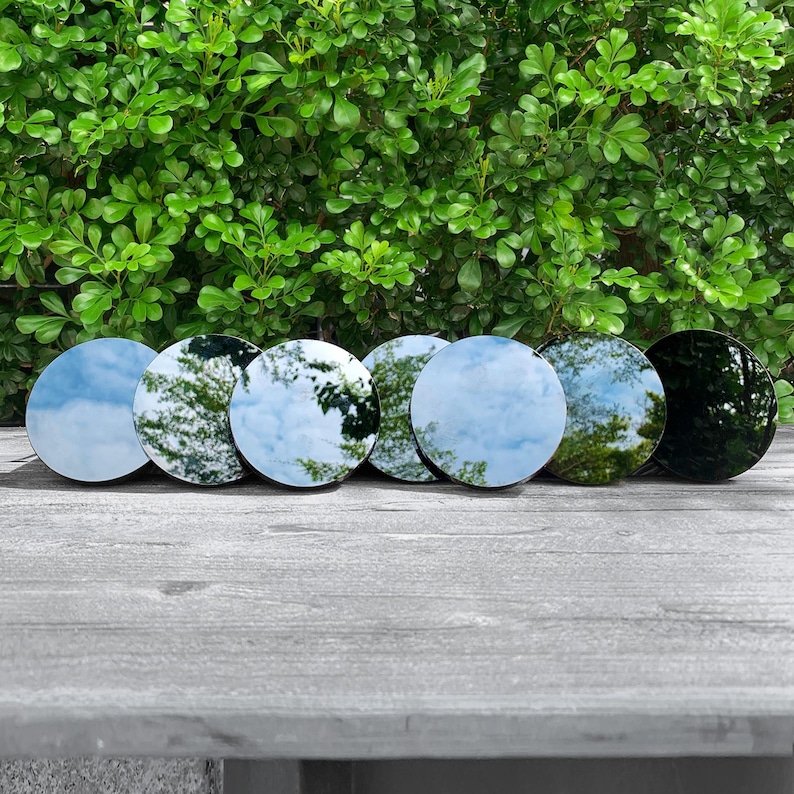May include: A row of seven circular, reflective glass discs arranged horizontally. Each disc features a smooth, polished surface that mirrors the surrounding environment, including the sky and foliage, with one disc appearing darker than the others.