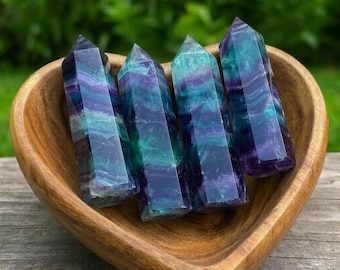 May include: Four polished fluorite crystal points with distinct bands of deep purple, teal, and translucent green. These hexagonal towers are arranged upright inside a smooth, heart-shaped wooden bowl with a visible natural grain pattern.