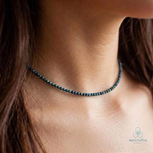 May include: A delicate beaded necklace featuring small, dark green and blue beads. The necklace is worn around the neck, highlighting the subtle colour variations. The beads are strung together to create a simple, elegant choker style.