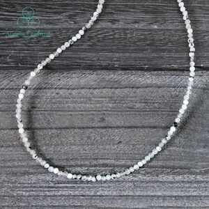 May include: Delicate necklace featuring small, round, white and gray beads. The beads are strung together to form a single strand. The necklace is displayed on a gray wooden surface. The text "Angelic Creations" is in the upper left corner.