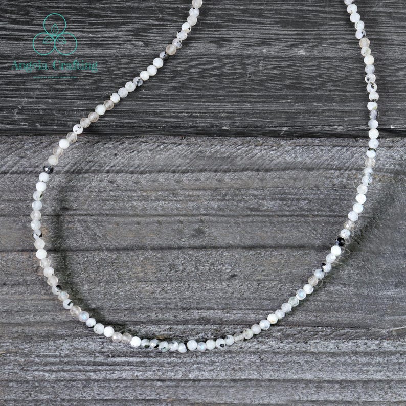 May include: Delicate necklace featuring small, round, white and gray beads. The beads are strung together to form a single strand. The necklace is displayed on a gray wooden surface. The text "Angelic Creations" is in the upper left corner.