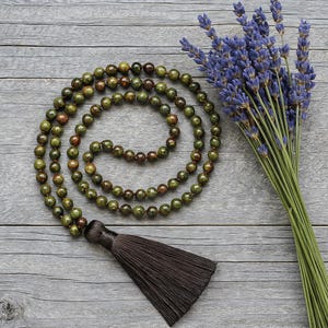 May include: A green and brown beaded mala necklace with a dark brown tassel. A bundle of purple lavender flowers rests beside the necklace on a gray wooden surface. The mala beads are round and vary in color.