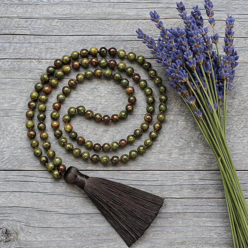 May include: A green and brown beaded mala necklace with a dark brown tassel. A bundle of purple lavender flowers rests beside the necklace on a gray wooden surface. The mala beads are round and vary in color.