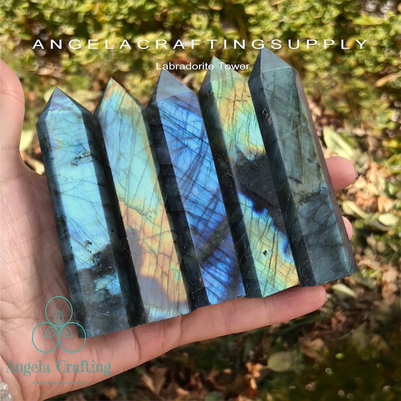 May include: Five polished labradorite towers with blue, green, and gold iridescent flashes are held in a hand. The towers are rectangular with pointed tops. The text "ANGELACRAFTINGSUPPLY" and "Labradorite Tower" are visible.