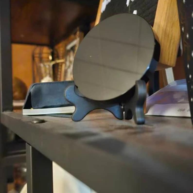 May include: A circular black mirror with a smooth, reflective surface is mounted on a black, four-legged stand. The mirror is positioned upright, displaying a dark, glossy finish that reflects the surrounding environment in a subtle, muted manner.