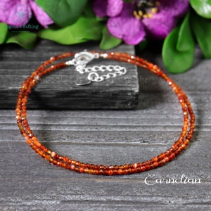 May include: A double-strand carnelian necklace with a silver clasp. The necklace is composed of small, faceted orange-red beads. The word "Carnelian" is written in white script at the bottom of the image. The necklace is displayed on a dark gray wooden surface.