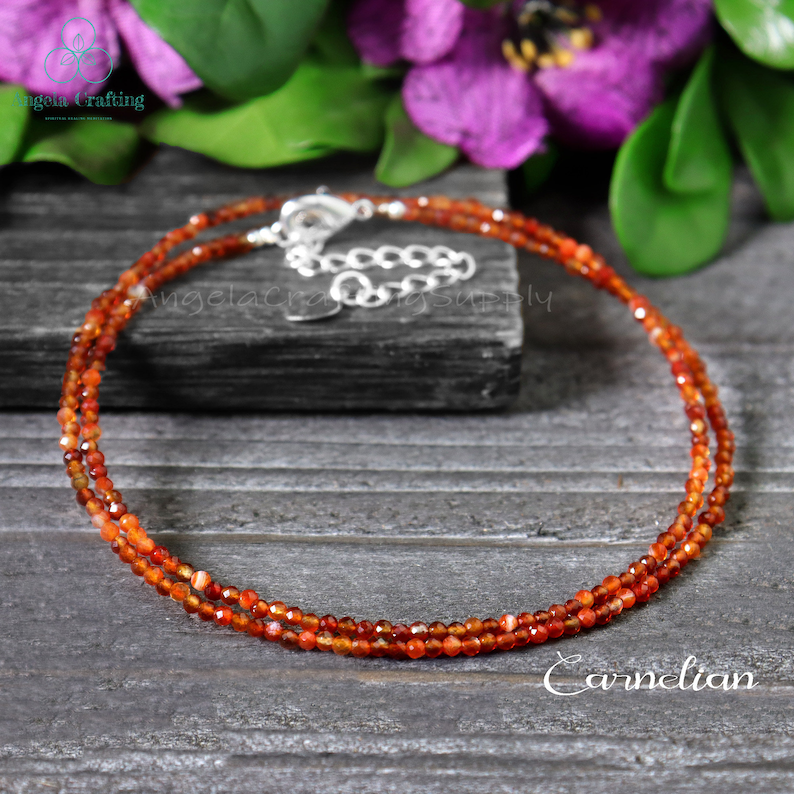 May include: A double-strand carnelian necklace with a silver clasp. The necklace is composed of small, faceted orange-red beads. The word "Carnelian" is written in white script at the bottom of the image. The necklace is displayed on a dark gray wooden surface.