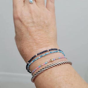May include: A stack of four bracelets consisting of one dark blue beaded bracelet, one light blue beaded bracelet, one multi-coloured pastel beaded bracelet with a central gold bead, and one thick silver-toned twisted metal cuff bracelet.