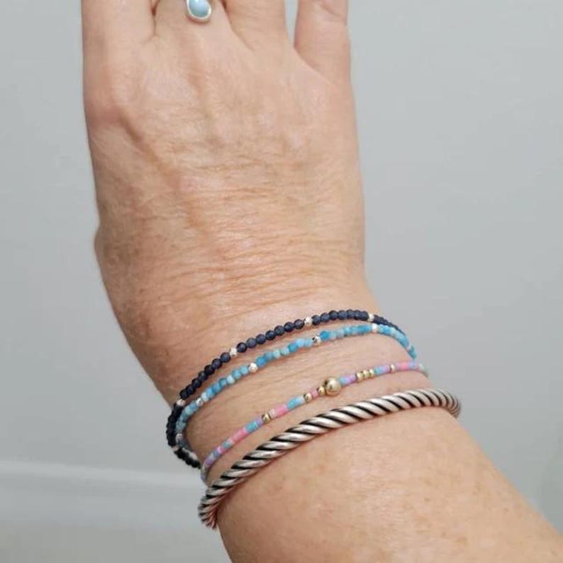 May include: A stack of four bracelets consisting of one dark blue beaded bracelet, one light blue beaded bracelet, one multi-coloured pastel beaded bracelet with a central gold bead, and one thick silver-toned twisted metal cuff bracelet.