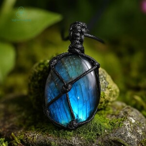 May include: A blue and green labradorite pendant, encased in a black woven macrame. The gemstone has a polished, oval shape and is suspended from a black cord. The background is a blurred view of green foliage and moss.