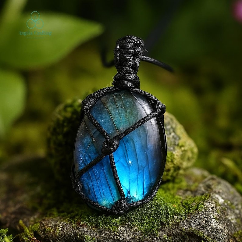 May include: A blue and green labradorite pendant, encased in a black woven macrame. The gemstone has a polished, oval shape and is suspended from a black cord. The background is a blurred view of green foliage and moss.