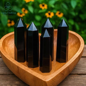 May include: Five polished black obsidian crystal points with hexagonal bases and pointed tops arranged inside a smooth, light-brown wooden bowl shaped like a heart. The crystals have a reflective, dark surface and are standing upright.