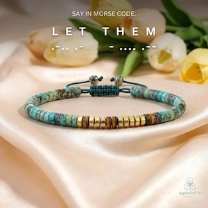 May include: A turquoise and gold beaded bracelet with the text "LET THEM" in Morse code. The bracelet features a sliding knot closure and is displayed on a satin surface with flowers in the background.