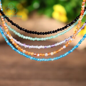 May include: A collection of delicate beaded necklaces in various colors, including black, green, purple, orange, and blue. Each necklace is composed of small, faceted beads, creating a sparkling effect. The necklaces are displayed against a blurred background.